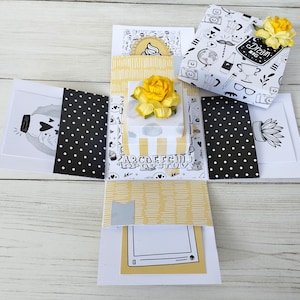 May include: An open, handmade explosion box with yellow and white patterned paper. The box features black and white illustrations, yellow paper accents, and yellow flower embellishments. The box has the words "Dream Big" on the top.
