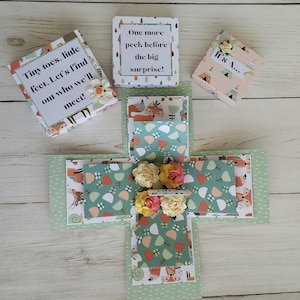 May include: A handmade explosion box with a baby shower theme. The box is decorated with floral and woodland animal patterns in shades of green, peach, and white. The box has several small boxes with text that reads "Tiny toes, little feet. Let's find out who we'll meet!" and "One more peek before the big surprise!"