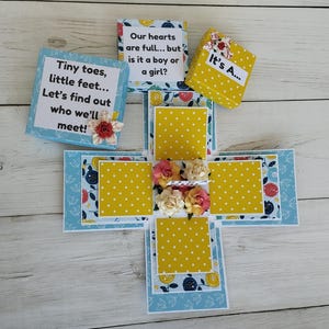 May include: A handmade gender reveal explosion box with blue and yellow accents. The box features text that reads "Tiny toes, little feet... Let's find out who we'll meet!" and "Our hearts are full... but is it a boy or a girl?"