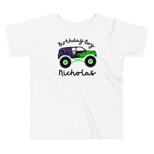 May include: White short-sleeved t-shirt with a green monster truck graphic with purple flames and a skull. The text "Birthday Boy Nicholas" is printed on the shirt.