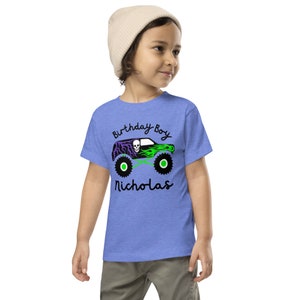 May include: A blue t-shirt with a green and purple monster truck graphic. The text "Birthday Boy Nicholas" is printed on the shirt.