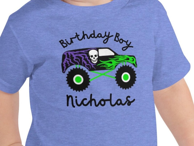 May include: A blue t-shirt with a green and purple monster truck graphic. The truck has a skull on the side and the text "Birthday Boy Nicholas" is printed on the shirt.