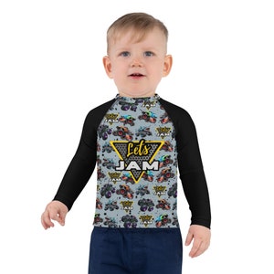 May include: A black and gray long-sleeved rash guard with a gray and black monster truck pattern. The shirt has a yellow triangle with the text "Let's Jam" in black and yellow letters.