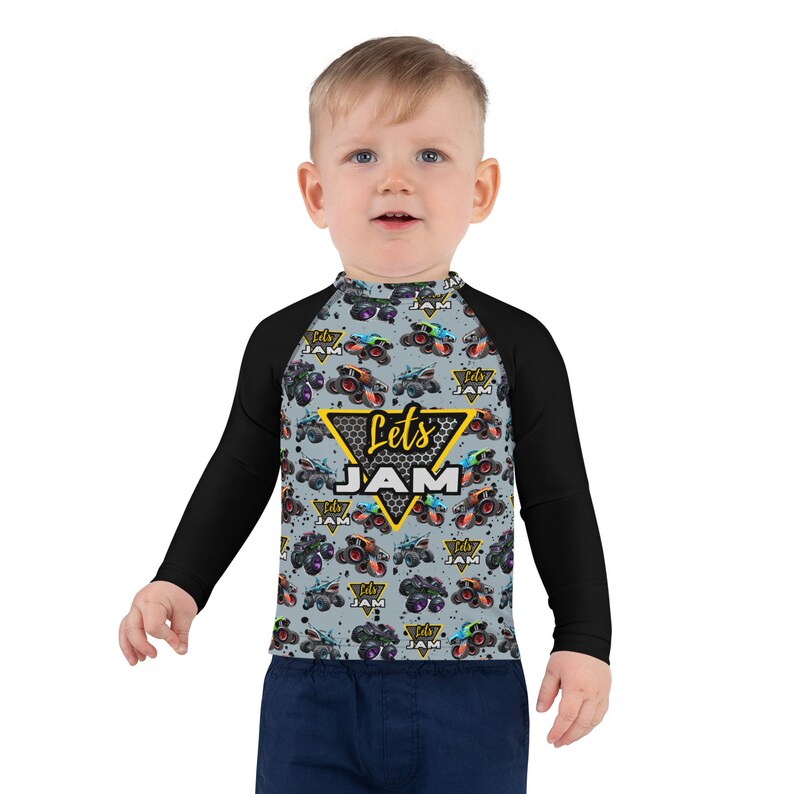 May include: A black and gray long-sleeved rash guard with a gray and black monster truck pattern. The shirt has a yellow triangle with the text "Let's Jam" in black and yellow letters.
