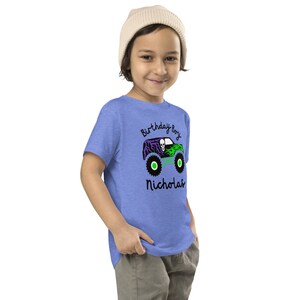 May include: A blue t-shirt with a green, purple, and black monster truck graphic. The text "Birthday Boy Nicholas" is printed on the shirt.