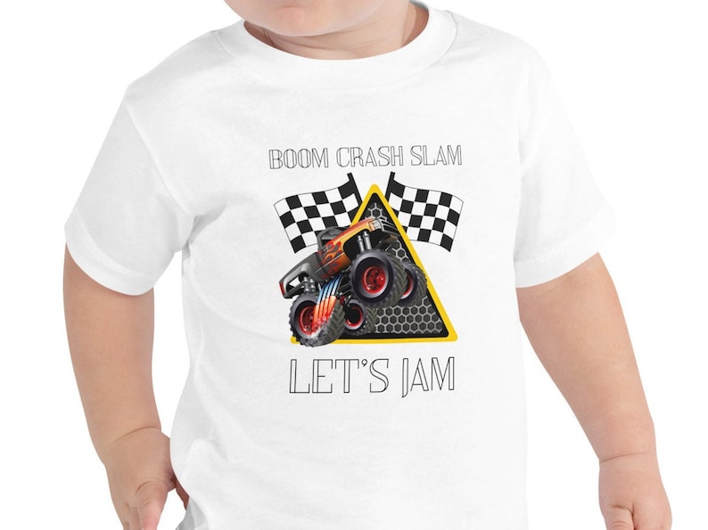 May include: White baby onesie with a black and orange monster truck graphic. The truck is driving through a yellow triangle with black checkered flags on either side. The text "BOOM CRASH SLAM LET'S JAM" is printed on the onesie.