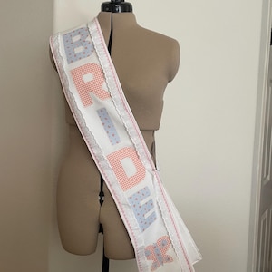 May include: White sash with the word "BRIDE" in patchwork letters. The letters are in red gingham and blue heart patterns. The sash is trimmed with white lace and pink stitching, and is displayed on a mannequin.