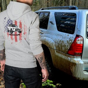 May include: A grey hoodie with a white and blue American flag design featuring a white SUV with the word "FREEDOM" printed below. The hoodie is being worn by a person standing next to a muddy silver SUV.