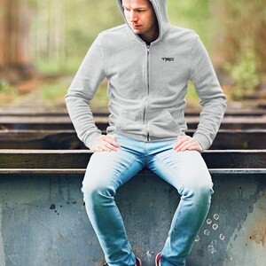 May include: A man wearing a grey hooded sweatshirt with the word "TRD" embroidered on the chest, blue jeans, and red trainers. He is sitting on a metal surface.