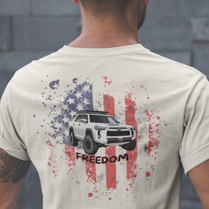 Puede incluir: Una camiseta blanca con un diseño de bandera estadounidense desgastado que presenta un SUV blanco y la palabra "FREEDOM" impresa en la espalda.