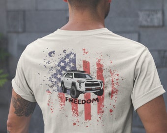 Op de afbeelding: Een wit T-shirt met een verweerd Amerikaans vlagontwerp met een witte SUV en het woord "FREEDOM" gedrukt op de achterkant.