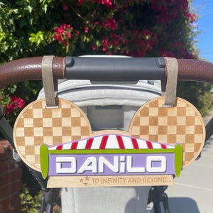 May include: A wooden stroller charm shaped like Mickey Mouse ears with a purple sign that says "DANILO" and "TO INFINITY AND BEYOND..." in white text. The sign has a red and white striped awning and green trim.