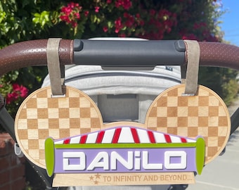 Puede incluir: Un amuleto de cochecito de madera con forma de orejas de Mickey Mouse con un letrero morado que dice "DANILO" y "TO INFINITY AND BEYOND..." en texto blanco. El letrero tiene un toldo a rayas rojo y blanco y un borde verde.