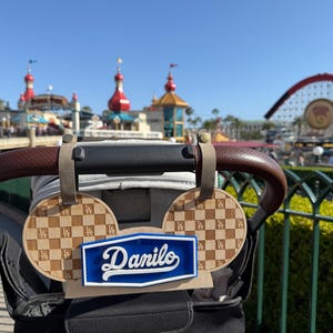 May include: A stroller accessory featuring a blue and white sign with the name "Danilo" and two wooden Mickey Mouse ear-shaped accents. The ears have a checkerboard pattern and the LA Dodgers logo. The accessory is attached to a stroller handle.