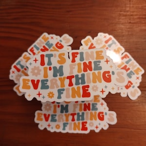 May include: A stack of white stickers with the text "It's fine, I'm fine, everything's fine" in colorful, retro-style lettering. The stickers feature a variety of small, colorful flowers and stars.