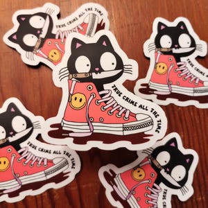 May include: A set of four black cat stickers with a pink and white sneaker design. Each sticker features a black cat with a knife in its mouth and the text "True Crime All The Time".