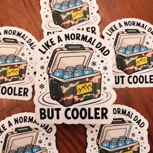 May include: A collection of stickers featuring a cooler design with the text "LIKE A NORMAL DAD BUT COOLER". The cooler is open, revealing blue cans inside, and has a camouflage pattern. Snowflakes and mountain graphics are also present.
