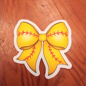 May include: A yellow bow-shaped sticker with red stitching details, resembling a softball. The sticker has a white border and is set against a wooden background. The bow is a bright, cheerful design.