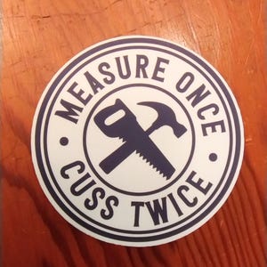 May include: A round sticker with a white background and a navy blue border. The text "MEASURE ONCE" and "CUSS TWICE" are in a circular pattern. A saw and hammer are crossed in the center.
