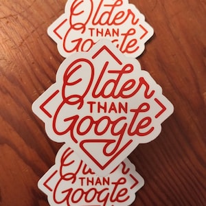 May include: Three white stickers with red text that reads "Older Than Google" in a stylized font. The stickers are diamond-shaped with a house-like top. The stickers are stacked on a wooden surface.