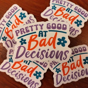 May include: A collection of white stickers with the phrase "Pretty Good at Bad Decisions" in purple, teal, and orange lettering. Each sticker has a floral accent. The stickers are arranged on a wooden surface.