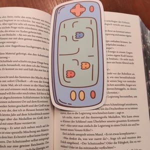May include: A sticker of a retro handheld video game console. The light blue console has a cross-shaped directional pad, buttons, and a screen displaying a maze-like game with three small character icons and three score indicators.