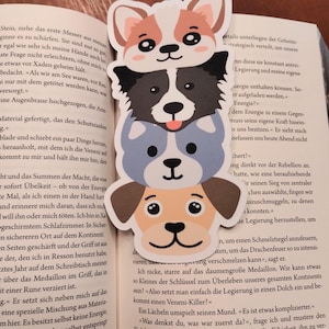 May include: A stack of four dog-shaped paper bookmarks. The top dog is a corgi, followed by a border collie, a blue-gray dog, and a tan dog. The bookmarks are on a book page.