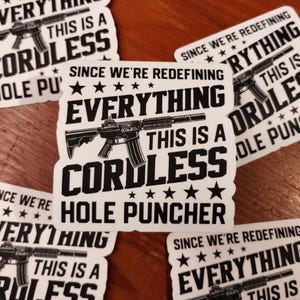 May include: White stickers with black text and a graphic of a rifle. The text reads "SINCE WE'RE REDEFINING EVERYTHING THIS IS A CORDLESS HOLE PUNCHER". The stickers are scattered on a wooden surface.