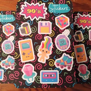 May include: A sheet of 90's themed stickers with a black background and colorful designs. The stickers include a cassette tape, a troll doll, a gameboy, roller skates, a ring with a gem, a floppy disk, a film roll, a tamagotchi, and a Rubik's cube.