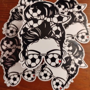 May include: A black and white sticker featuring a woman with a messy bun, wearing sunglasses shaped like soccer balls. The bun is decorated with a soccer ball patterned headband.