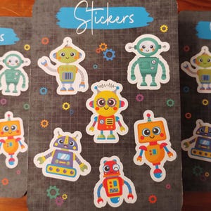 May include: A sheet of stickers featuring nine colorful robot designs. The robots are all different shapes and sizes, with various facial expressions and details. The stickers are printed on a black background with a blue banner at the top that says "Stickers".