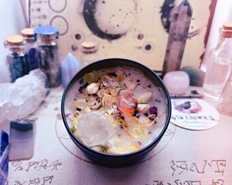 May include: A black candle jar with a white wax base and various crystals and dried flowers embedded in the wax. The candle is surrounded by other crystals and small glass bottles.