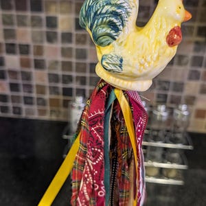 May include: A ceramic rooster ornament featuring a yellow body with blue-green tail feathers and a red comb. Below the rooster hangs a decorative tassel made of various fabric strips, including red plaid, solid yellow ribbon, and dark patterned textiles.