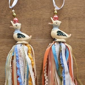 Puede incluir: Dos adornos decorativos de pato con cintas blancas para colgar. Cada pato tiene un cuerpo color crema, un cuello azul y una borla colorida de cintas e hilo en tonos naranja, azul y crema. Sobre una superficie de madera.