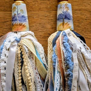 May include: Two decorative tassels with painted ceramic tops featuring a landscape scene. The tassels have a mix of ribbons, lace, and fabric in shades of blue, cream, and peach, with a wooden bead and gold accents.