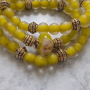 May include: A set of three stretch bracelets featuring frosted yellow glass beads and ornate gold-toned metal spacers. The center bracelet includes a larger, smooth yellow glass bead with a subtle white floral pattern at its core.