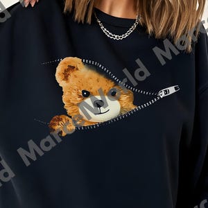 Puede incluir: Sudadera negra con un oso de peluche marrón asomando por un bolsillo con cremallera. El oso tiene una expresión amigable. La sudadera es de color negro liso, con mangas largas. El diseño es juguetón y caprichoso.