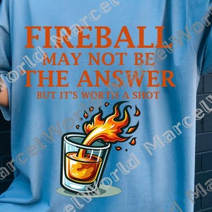 Op de afbeelding: Lichtblauw t-shirt met de oranje tekst "FIREBALL MAY NOT BE THE ANSWER BUT IT'S WORTH A SHOT". Het shirt heeft een afbeelding van een shotglaasje met vlammen. Een casual kledingstuk.