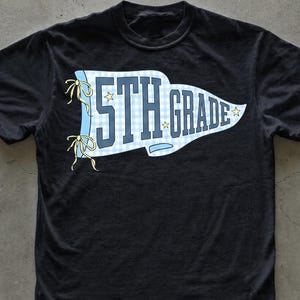 May include: A black short-sleeve t-shirt featuring a light blue and white gingham pennant graphic on the chest. The pennant contains the dark blue text 5TH GRADE, accented with small yellow stars and yellow ribbons tied on the left side.