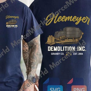 May include: Navy blue t-shirt with a front pocket design featuring the name "Heemeyer" and a bulldozer graphic. The back of the shirt displays the name "Heemeyer" in gold script above a bulldozer graphic and the text "DEMOLITION INC. GRANBY CO. EST. 2004."