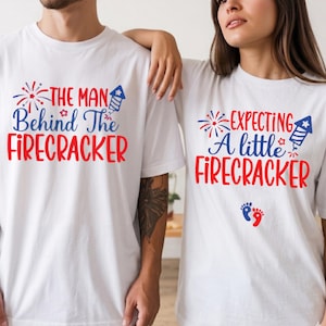May include: Two white t-shirts with red and blue text. One shirt says "THE MAN Behind The FIRECRACKER" and the other says "EXPECTING A little FIRECRACKER" with baby footprints. The shirts have a festive, patriotic theme.