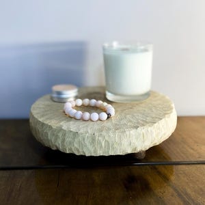以下が含まれることがあります： 木製の丸い、明るい色のトレイ。ガラスに入った白いキャンドル、小さな銀色の缶、ピンクのビーズブレスレットが置かれています。トレイは濃い木製の表面に置かれています。