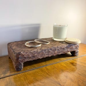 以下が含まれることがあります： 濃い茶色の彫刻が施された木製のプラットフォーム。2つのシルバーブレスレット、透明なガラスのキャンドル、木製の蓋が置かれています。プラットフォームは長方形で、表面は質感があり、短い脚が付いています。キャンドルは白いワックスと木の芯です。