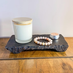 以下が含まれることがあります： 木製の蓋が付いた透明なガラス瓶に入ったキャンドルが、暗い彫刻が施された木製のトレイの上に置かれています。 ピンクと白の石のビーズブレスレットと小さな金属製の缶もトレイにあります。