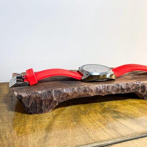 以下が含まれることがあります： 赤色のシリコンバンドが付いたシルバーの時計が、濃い茶色の彫刻が施された木製のディスプレイに置かれています。 時計の文字盤が見え、バンドはシルバーの留め金で固定されています。 木製ディスプレイは質感のある表面です。