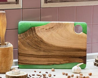 May include: A kitchen countertop scene featuring a wooden cutting board with green resin, a wooden utensil holder with a spatula, and a white teapot with a cup. Decorative items include a small astronaut figurine and a garlic bulb.