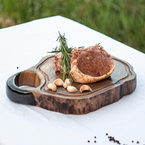 May include: A rustic wooden serving board with a dark handle and a natural edge. The board holds a cooked meat dish, sprigs of rosemary, and several cloves of garlic. Black peppercorns are scattered around the board.