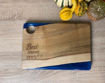 Peut inclure: Planche à découper en bois avec un accent en résine bleue et le texte gravé "Best mom ever!". Un trou est percé dans le coin supérieur gauche. Des courges de différentes couleurs sont en arrière-plan. La planche est de forme rectangulaire.