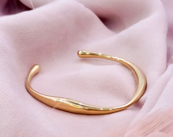 May include: A gold-toned, open-ended bangle bracelet with a smooth, curved design. The bracelet is lying on a pink fabric background.