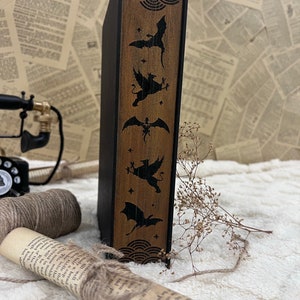 Pode incluir: Um livro preto com uma lombada de madeira que apresenta um padr&atilde;o repetitivo de morcegos estilizados e outras criaturas. A lombada &eacute; decorada com um padr&atilde;o de gr&atilde;o de madeira.